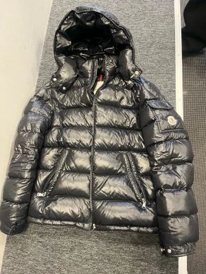 Moncler Maya Jacka svart - Säljer en svart pufferjacka från Moncler med glansig finish, stor huva och dragkedja framtill. Jackan har två snedställda fickor med dragkedja och en extra ficka med knapp på ärmen där Moncler-loggan sitter. Perfekt för kalla dagar och riktigt snygg streetstil.