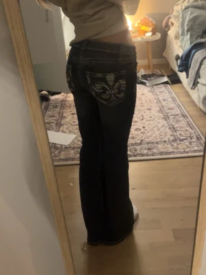 Mörkblå bootcut jeans från Gina Tricot - Snygga mörkblå jeans från Gina Tricot med bootcut-modell och kontrastsömmar. Bakfickorna har broderad detalj i vitt och silver, vilket ger en unik look. Jeansen har klassisk knapp och dragkedja framtill.