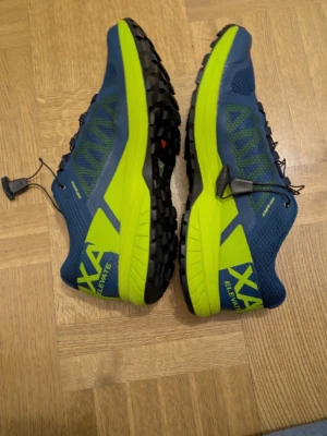 Salomon XA Elevate blå/gula sneakers - Säljer ett par Salomon XA Elevate sneakers i blått och neon-gult. Skorna har grov sula för extra grepp, snabbsnörning och mesh-överdel för bra ventilation. Perfekta för dig som gillar att springa i terräng eller bara vill ha riktigt snygga och sportiga skor.