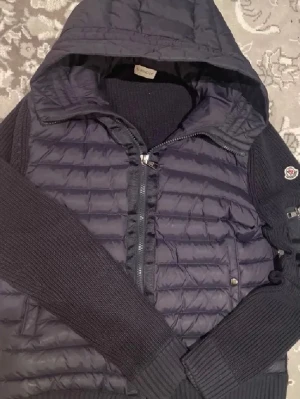 Mörkblå pufferjacka från Moncler - Säljer en mörkblå pufferjacka från Moncler med stickade ärmar och huva. Jackan har dragkedja framtill, quiltad kropp och ribbade muddar. Klassisk Moncler-logga på ärmen och snygga detaljer som ger en sportig vibe.