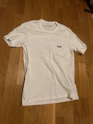 Vit t-shirt från BOSS - Säljer en clean vit t-shirt från BOSS med diskret logga på bröstet. Klassisk rund hals och korta ärmar. Perfekt basic-plagg i mjuk bomull som funkar till allt. Enkel och stilren design.
