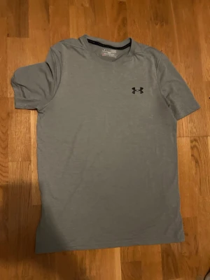 Grå t-shirt från Under Armour - Säljer en enkel och stilren grå t-shirt från Under Armour med svart logga på bröstet. T-shirten har korta ärmar och är gjord i ett mjukt och lätt material som passar perfekt till träning eller chill.