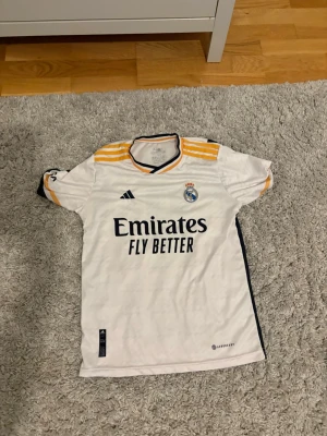 Real Madrid  - Säljer en vit Real Madrid matchtröja från Adidas med gula och svarta detaljer. Tröjan har klubbmärke, sponsortryck och nummer 9 med Benzema på ryggen. Tillverkad i lätt polyester med klassisk passform och Aeroready-teknologi.
