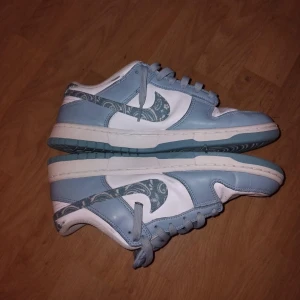 Nike Dunk Low blå/vit paisley - Nike Dunk Low sneakers i ljusblått och vitt med paisleymönstrad swoosh och häl. Skorna har klassisk snörning, perforerad tåbox och platt sula. Tillverkade i skinn och textil för en clean och fräsch look. Perfekta för dig som gillar unika detaljer.