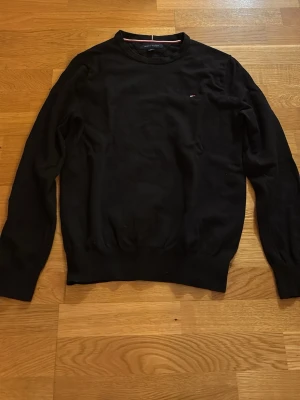 Svart tröja från Tommy Hilfiger - Klassisk svart tröja från Tommy Hilfiger med rund halsringning och diskret logga på bröstet. Tröjan har långa ärmar och ribbade muddar vid ärmslut och nederkant. Perfekt till höst och vinter, enkel att matcha med allt.
