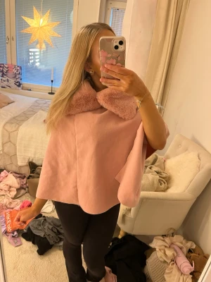 Rosa kappa med fuskpäls-krage - Superfin ljusrosa kappa med bred, fluffig fuskpäls-krage. Modellen är loose och har en cape-liknande form utan synliga knappar eller dragkedja. Perfekt för dig som vill ha en söt och trendig look med extra mysig känsla.