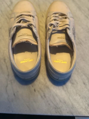 Saint Laurent vita sneakers med snörning - Säljer ett par vita Saint Laurent sneakers med handskriven logga på sidan. Skorna har rund tå, platt sula och snörning framtill. Ovandelen är i canvas med detaljer i skinn och insidan är ljusbeige. Perfekt för dig som gillar stilrena och exklusiva sneakers. På grund av fläcken är skorna 1000kr billigare än vanligt försäljningspris pris på saint laurents skor i detta skick 