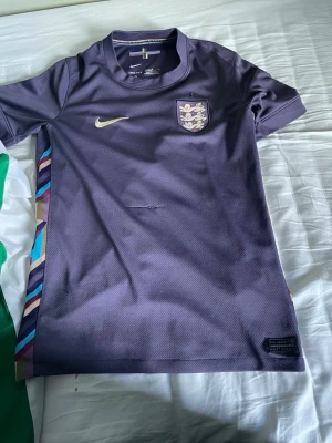 Stilig Englands tröja - En riktig Englands tröja från Vm 2022 den är en mörk lila ton med guldigt märke och stiligt mönster på sidorna PS Tröjan har en söm som är avdragen på framsidan men bortsett ifrån det så är den i bra skick.