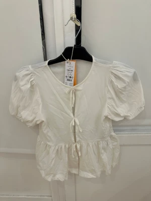 Vit blus med puffärm från Gina Tricot - Supersöt vit blus från Gina Tricot med puffiga korta ärmar och tre knytband framtill. Blusen har en volangkant nedtill och är gjord i mjuk bomull. Perfekt för dig som gillar romantiska detaljer och en luftig känsla.