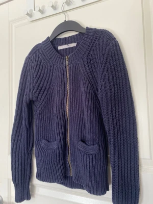Mörkblå stickad kofta Tiger of Sweden - Mörkblå, chunky stickad kofta från Tiger of Sweden i storlek S. Koftan har grov ribbstickning, dragkedja framtill och två små fickor. Tillverkad i 100% bomull och har lång ärm. Perfekt för lager-på-lager under kyliga dagar.