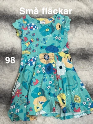 Turkos blommig barnklänning stl 98 - Färgglad turkos klänning från Polarn o pyret med korta ärmar och ett lekfullt mönster av blommor, fiskar och sköldpaddor i blått, gult, vitt och rött. har en snurrig kjol som är perfekt för lek.🌸 Små fläckar. Storlek 98