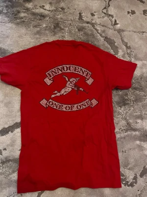 Röd t-shirt med strasstenar från One of One - Röd t-shirt med kort ärm från One of One. Baksidan har ett stort tryck med texten 'INNOCENT' och 'ONE OF ONE' i strasstenar samt en ängel i mitten. T-shirten är i bomull och har en klassisk passform. Perfekt för dig som vill sticka ut med detaljer.