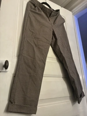 Rutig kostymbyxa Holly & Whyte - Snygga kostymbyxor från Holly & Whyte i klassiskt rutigt mönster i beige, brunt och svart. Byxorna har raka ben, bälteshällor och två bakfickor. Perfekta för en stilren look och passar till både sneakers och boots.