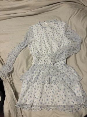 Blommig kortklänning från Gina Tricot 158/164 - Supersöt vit kortklänning från Gina Tricot med blått blommönster. Klänningen har långärmade, transparenta ärmar, smock i midjan och volangdetaljer på kjolen. Perfekt för sommarens alla tillfällen!