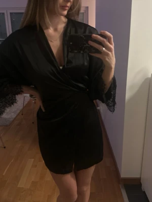 Svart kimono med spetsdetaljer - Säljer en svart kimono i sidenliknande material med breda ärmar och snygga spetsdetaljer vid ärmsluten. Den har en elegant och flowy passform som ger en lyxig känsla. 