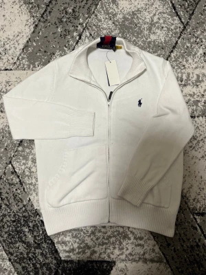 Vit kofta från Polo Ralph Lauren - Vit stickad kofta från Polo Ralph Lauren med dragkedja framtill och ribbade muddar. Klassisk broderad logga i marinblått på bröstet och ståkrage med snygga röd- och marinblå detaljer i nacken. Perfekt till lager på lager under höst och vinter.