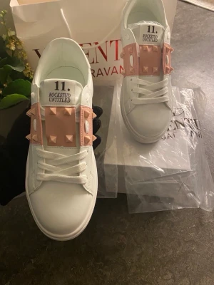 Valentino Garavani Skor - Säljer ett par vita Valentino Rockstud Untitled sneakers med ljusrosa detaljer och ikoniska nitar. Skorna har snörning, rund tå och platt sula med mönstrad undersida. Tillverkade i skinn och har en chunky look med coola rockstuds längs sidan.