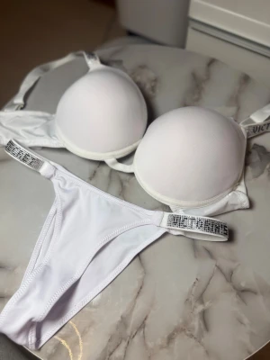 Vit push-up bh från Victoria's Secret - Säljer en vit push-up bh från Victoria's Secret med vadderade kupor och justerbara axelband. Axelbanden har glittriga detaljer med små stenar som ger en extra lyxig känsla. Perfekt för dig som vill ha något snyggt och bekvämt under kläderna.
