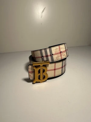 Rutig Burberry bälte med guldspänne - Snyggt Burberry bälte med klassiskt rutigt mönster i beige, svart och rött på ena sidan och svart med diskret rutmönster och logotyp på andra sidan. Bältet har ett stort guldigt B-spänne som ger en lyxig touch. Materialet är skinn med präglad yta.