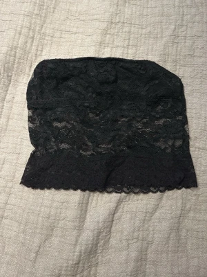 Svart spets bandeau tubtopp - Säljer en svart bandeau tubtopp med vacker spets och mjukt foder på insidan. Toppen är axelbandslös och har en feminin scallop-kant nedtill. Perfekt att styla med jeans eller kjol för en trendig look.