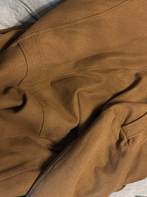 Brun rock från Indigo, storlek 2XL - Säljer en brun rock från Indigo i storlek 2XL. Jackan har klassisk krage, svarta knappar framtill och slät yta. Fodrad med svart tyg på insidan och har fickor med lock. Perfekt för dig som gillar stilrena och tidlösa plagg.