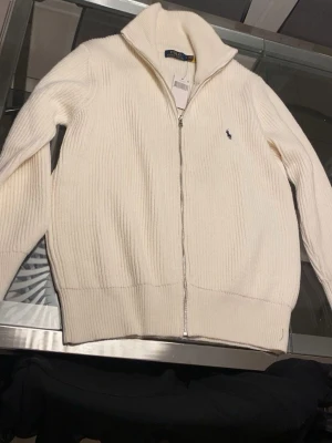 Vit stickad kofta från Polo Ralph Lauren - Säljer en vit stickad kofta från Polo Ralph Lauren i storlek M. Koftan har ribbad struktur, lång ärm och silverfärgad dragkedja framtill. Klassisk krage och den ikoniska Polo-loggan broderad på bröstet. Perfekt för lager-på-lager under kyliga dagar. Pris går o diskutera bara o höra av sig 