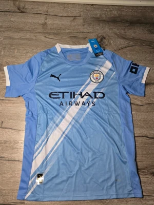 Manchester City fotbollströja - Manchester Citys officiella matchtröja från Puma i ljusblått med vita detaljer och klubbmärke på bröstet. Tröjan har rund hals, korta ärmar och diagonal randig design framtill. Materialet är lätt polyester som andas och passar perfekt för fotboll.