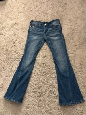 True Religion Joey Low Rise Flare Jeans - Säljer ett par blå jeans från True Religion, modell Joey Low Rise Flare. De har låg midja, utställda ben och klassiska fickor med dekorativa sömmar och silverfärgade knappar. Materialet är jeans med snygga slitningar och tvättad look.