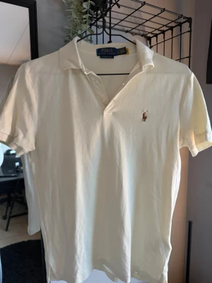 Gul/vit pikétröja från Polo Ralph Lauren - Säljer en klassisk vit/guk pikétröja från Polo Ralph Lauren i slim fit. Tröjan har korta ärmar, krage och knappar framtill samt den ikoniska broderade loggan på bröstet. Perfekt för en clean och stilren look.