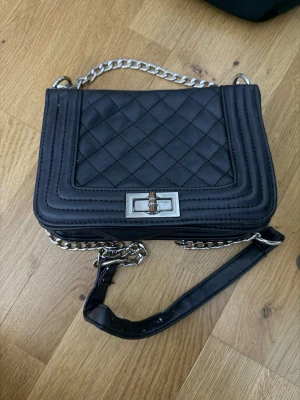 Svart quiltad axelväska med kedja - Snygg svart axelväska med quiltad design och silverfärgad kedja som axelrem. Väskan har ett rektangulärt spänne i metall framtill och är tillverkad i skinn. Perfekt storlek för det viktigaste och ger en cool vibe till din outfit.