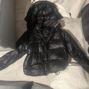 Burberry vinterjacka - Säljer en svart pufferjacka från Burberry med huva och stora fickor framtill. Jackan har en glansig finish, dragkedja och ribbade muddar vid ärmsluten. Perfekt för kalla dagar och har en riktigt schysst streetstil. Hör av dig om du vill veta mer! 