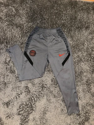 Grå Nike PSG träningsbyxor - Snygga grå träningsbyxor från Nike med Paris Saint-Germain-logga i svart och rött på ena benet. Byxorna har röd Nike Swoosh, svarta detaljer längs benen och är tillverkade i ett mjukt syntetmaterial. Perfekta för dig som gillar fotboll och streetwear.