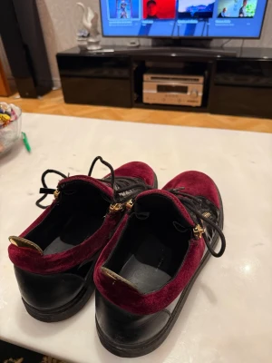 Giuseppe Zanotti vinröda - Säljer ett par unika sneakers i vinröd sammet med svarta skinnpaneler och svarta snören. Skorna har guldfärgade dragkedjor på sidorna och svarta detaljer. Snygg platt sula och rund tå, perfekt för dig som vill sticka ut med din stil.