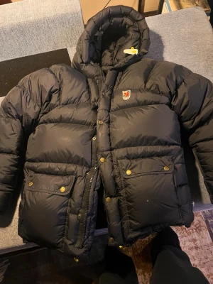 Svart pufferjacka från Fjällräven - Säljer en svart pufferjacka från Fjällräven i storlek S. Jackan har huva, stora fickor med guldfärgade knappar och dragkedja framtill. Perfekt för kalla dagar och har den klassiska Fjällräven-loggan på bröstet. Jackan är riktigt fluffig och har en schysst oversized känsla.