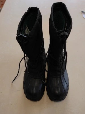 Svarta vinterstövlar från Blaxter - Svarta vinterstövlar från Blaxter med grov mönstrad sula och högt skaft. Skorna har snörning framtill och är tillverkade i slitstark syntet och skinn. Insidan är fodrad för extra värme och komfort under kalla dagar. Perfekt för snö och vinterväder.