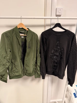 Olivgrön bomberjacka med dragkedja - Säljer en olivgrön bomberjacka med klassisk ribbad krage och muddar. Jackan har dragkedja framtill, fickor med knapp och är tillverkad i ett lätt, glansigt material. Perfekt för dig som vill ha en stilren och enkel jacka med streetkänsla.