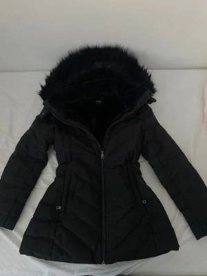 Svart vinterjacka med fuskpäls från Zara - Svart pufferjacka från Zara med fluffig fuskpäls på huvan och quiltad design. Har haft på mig den en sesången så den är i mycket bra skick och pälsen är mjuk o fluffig som helt ny. Jackan har dragkedja framtill, figurnära passform och långa ärmar. Perfekt för kalla dagar och har snygga detaljer vid midjan.