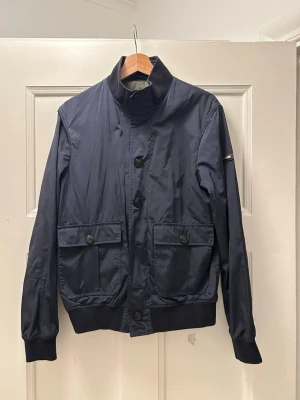 Mörkblå Jack från Hackett London - Säljer en mörkblå Jack från Hackett London i storlek S. Jackan har hög krage, stora fickor med knappar framtill och ribbade muddar. Tillverkad i polyester och har en clean, stilren look som passar perfekt till vardagsoutfits.