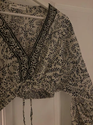 Mönstrad blus från Pull&Bear - Säljer en superfin blus från Pull&Bear med blått och vitt bladmönster. Blusen har v-ringning med broderade detaljer, vida ärmar och knytband med tofsar i midjan. Croppad modell som ger en avslappnad och bohemisk vibe.