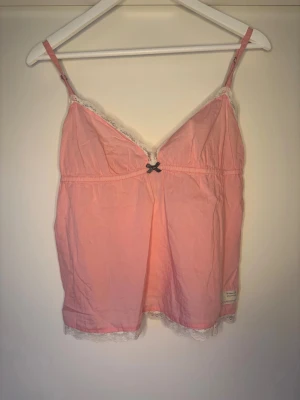 Rosa Odd Molly linnetopp med spets - Supersöt rosa linnetopp från Odd Molly med justerbara axelband, vit spetskant vid halsringning och nederkant samt en liten rosett framtill. Broderad Odd Molly-logga på ryggen och tygmärke vid nederkant. Perfekt för dig som gillar romantisk och feminin stil.