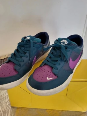 Nike SB sneakers blå/lila 42 - Nike SB sneakers i blått och lila med klassisk vit sula och lila swoosh på sidan. Skorna har snörning och perforerad tå för extra ventilation. Låg modell med rund tå och platt sula, tillverkade i syntetmaterial och skinn. Perfekt för dig som gillar färgstarka sneakers.