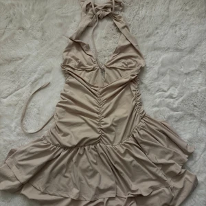 Beige omlott kortklänning med volang - Supersöt beige kortklänning med omlott och knyt i nacken. Klänningen har volang i nederdelen och rynkade detaljer framtill. Djup v-ringning och ärmlös design ger en trendig  look. Perfekt för varma dagar och sommarhäng.