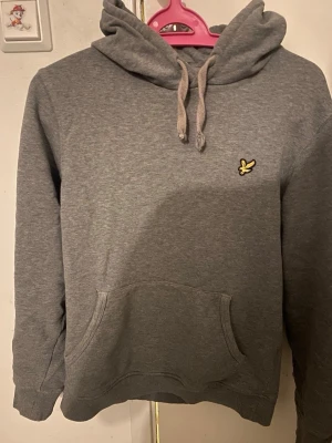 Grå lyle & Scott hoodie - Grå hoodie i klassisk modell Stilren grå hoodie i mjukt och skönt material. Tröjan har huva med dragsnören, känguruficka framtill och ribbade muddar vid ärmslut och nederkant. På bröstet finns en diskret broderad logotyp som ger ett sportigt och tidlöst intryck. Hoodien är i gott skick och passar perfekt för både vardag och fritid.