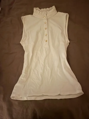 Vit ärmlös topp från Holly & Whyte - Säljer en vit ärmlös topp från Holly & Whyte by Lindex i storlek XS. Toppen har en hög krage med volangdetalj och fem dekorativa knappar framtill. Materialet känns mjukt och lätt, perfekt för varmare dagar. Snygg och stilren design som passar till många outfits.