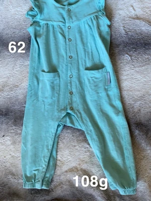 Turkos byxdress med fickor, stl 62 - Turkos byxdress med två praktiska fickor framtill och tryckknappar längs hela framsidan. Byxdressen har resår vid bensluten och är ärmlös, perfekt för en chill och bekväm stil.🌸 Fint använt skick. Storlek 62