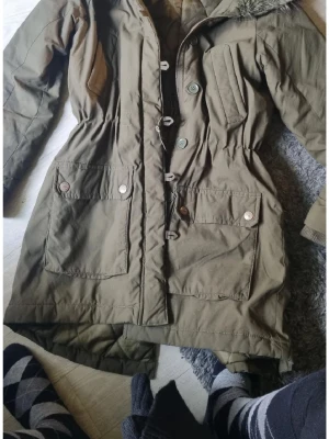 Olivgrön jacka med knappar och huva - Säljer en olivgrön jacka med stora fickor, knappar och dragkedja framtill. Jackan har huva med fuskpäls, markerad midja och långa ärmar. Perfekt för kyliga dagar och riktigt skön att bära. Passar dig som gillar en avslappnad men ändå snygg stil.