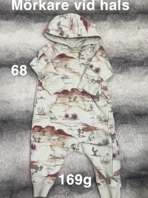 Beige mönstrad babyoverall från Newbie - Säljer en beige overall med huva från Newbie, dekorerad med ökeninspirerat mönster i bruna och grå toner. Overallen har tryckknappar vid halsen och mjuka muddar vid ärmslut och benslut. Perfekt för dig som gillar naturmotiv och bekväma plagg.🌸 Något mörkare vid halsen. Storlek 68