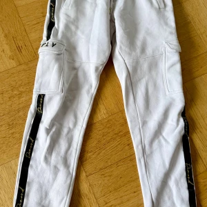 Vita cargopants Supply & Demand XS - Säljer ett par vita cargopants från Supply & Demand i storlek XS. Byxorna har svarta detaljer längs benen med guldfärgad text, stora fickor på sidorna och broderad logga på ficklocket. Tillverkade i mjukt bomullsmaterial med ribbad midja för extra komfort.