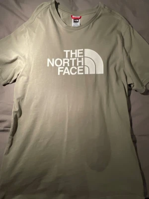 Olivgrön t-shirt från The North Face - Snygg olivgrön t-shirt från The North Face med stort vitt logotryck på bröstet och mindre logga på ryggen. Klassisk rund hals och korta ärmar. Tillverkad i mjuk bomull som känns skön mot huden. Perfekt för chill dagar eller när du vill ha en clean streetstyle.