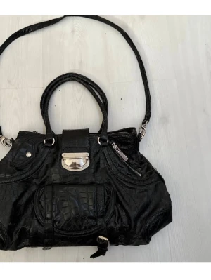 Svart vintage väska från Charmant - Säljer en svart vintage axelväska i större modell  från Charmant of Sweden i fejkskinn med croco-mönster. Väskan har både handtag och lång axelrem, flera fack med dragkedja och snygga silverdetaljer. Perfekt för dig som vill ha plats för allt och ändå se stylish ut. 